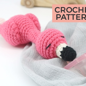 May include: Pink crocheted flamingo toy with a black eye and beak, attached to a clear pacifier. The toy is lying on a gray blanket. CROCHET PATTERN text is visible in the image.