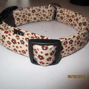 May include: A beige dog collar with a brown and green floral pattern. The collar has a black plastic buckle and a black metal D-ring.
