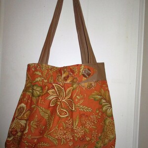May include: A large orange and brown tote bag with a floral pattern. The bag has two long brown straps and a brown leather flap with a button closure.