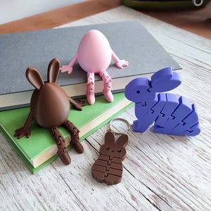 May include: A collection of Easter-themed 3D-printed objects. Includes a pink egg figure with arms and legs, a brown egg-shaped bunny, a purple segmented bunny, and a brown bunny keychain. The objects are arranged on books and a wooden surface.