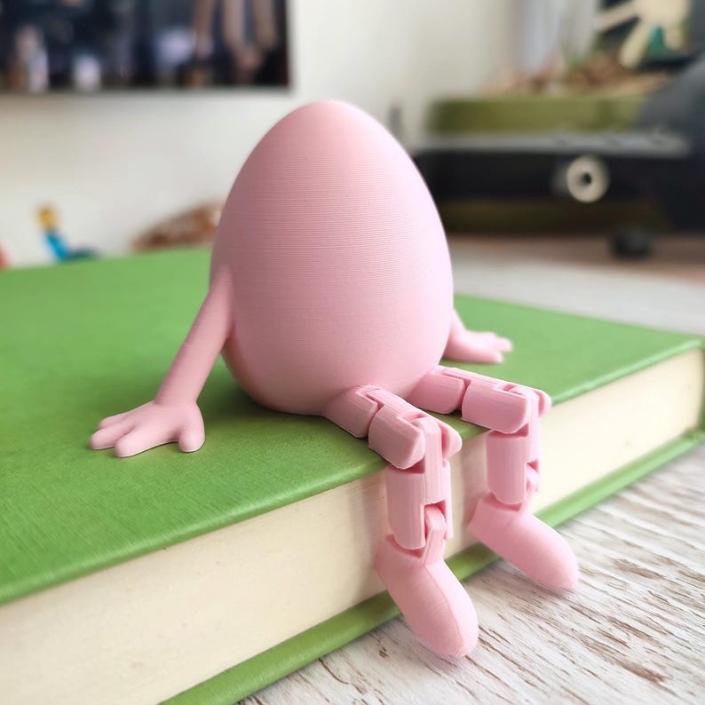 May include: A pink egg-shaped figurine with articulated arms and legs, sitting on a green book. The figurine's limbs are posed as if it is relaxing. The book has a green cover and white pages. The figurine is a whimsical decorative item.