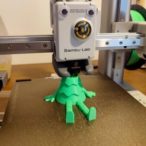May include: A green 3D-printed figure with arms and legs is on a textured plate. The figure is under a white device with the text "Bambu Lab" and "CAUTION HOT". The device has googly eyes and the text "move this cover for attend replacement".