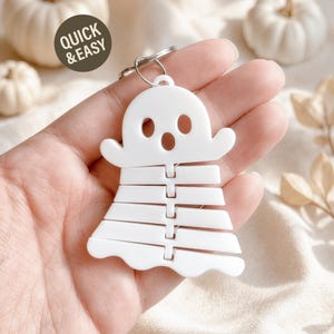 May include: A white ghost-shaped keychain featuring a segmented, articulated body design. The ghost has two oval eyes and a circular mouth cutout. A metal split ring is attached to the top. The text QUICK & EASY is printed on a dark circle.