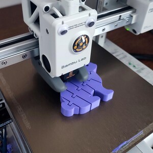 Puede incluir: Primer plano de una impresora 3D imprimiendo un objeto de pl&aacute;stico morado con forma de pez. La impresora es un modelo Bambu Lab y tiene un cuerpo blanco con un logotipo negro y naranja. El texto "remove this cover for stand replacement" es visible en la impresora.