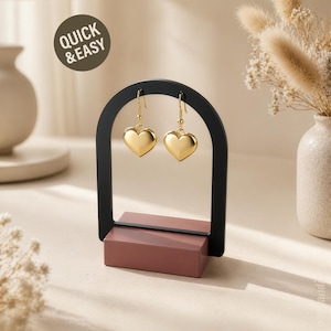 Puede incluir: Un expositor de pendientes negro con dos pendientes dorados en forma de corazón. El expositor tiene una base rectangular y una parte superior arqueada. El texto "QUICK & EASY" está en un círculo en la esquina superior izquierda.
