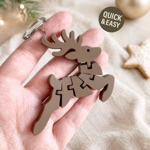 Pode incluir: Um porta-chaves em forma de rena castanha feito de peças de puzzle interligadas. O design apresenta uma rena a saltar com hastes e um anel metálico preso no topo. Um gráfico circular no canto exibe o texto QUICK & EASY.