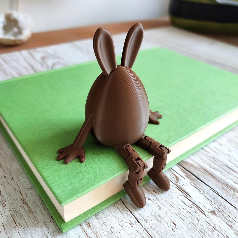 May include: A brown Easter bunny-shaped bookend with long ears and articulated legs, sitting on a green book. The bunny is a solid color and has a smooth texture. The book has a green cover and white pages.