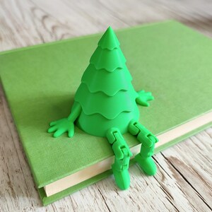 May include: A bright green, 3D-printed tree-shaped book holder with articulated arms and legs, sitting on a green book. The tree has a layered design and is resting on the book's cover. The book is closed and has a green cover.