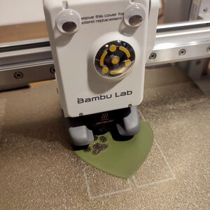 May include: A white 3D printer with googly eyes and the text "Bambu Lab" is printing a green heart-shaped object with a paw print design. The printer has a warning label that says "Caution Hot".