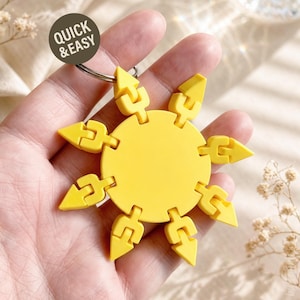 May include: A bright yellow sun-shaped keychain featuring a central circular disc surrounded by eight articulated, arrow-shaped points connected by hinges. A circular metal ring is attached to the top. A dark circular tag displays the text QUICK & EASY.