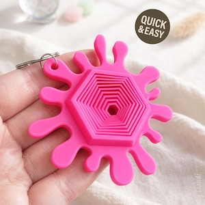 May include: A bright pink keychain featuring a unique, abstract design. The central element is a series of nested hexagons, surrounded by a splash-like outline. The words "QUICK & EASY" are printed on a brown circle.