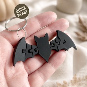 May include: A black articulated bat keychain featuring a segmented wing design and a silver metal split ring. A circular brown tag with the white text QUICK & EASY is attached to the top of the keychain, which is held in a palm.
