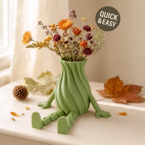 Può includere: Un vaso verde chiaro con corpo a spirale scanalato e braccia e gambe articolate, riempito con un bouquet di fiori secchi arancioni, gialli e rosso scuro. È visibile un adesivo marrone scuro con il testo bianco QUICK & EASY.