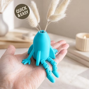 Peut inclure: Un vase turquoise en forme de fusée avec des jambes et des bras articulés, contenant des fleurs séchées. Le vase a un corps cylindrique avec des ailes en forme d'aileron. Le texte "QUICK & EASY" est dans un cercle en haut à gauche.