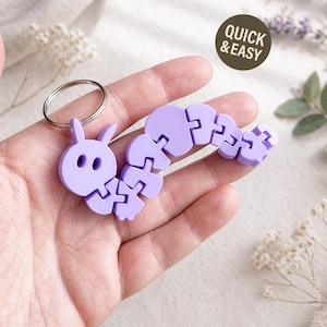 Puede incluir: Un llavero con forma de oruga de color lavanda. El llavero tiene un cuerpo segmentado y una cabeza con dos ojos redondos y dos orejas pequeñas. Un llavero de metal está unido a la cabeza. El texto "QUICK & EASY" está en un círculo marrón en el fondo.