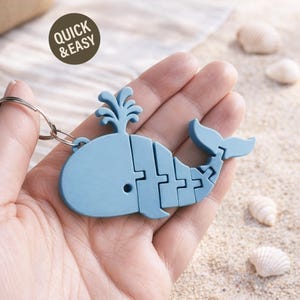 Puede incluir: Un llavero azul claro con forma de ballena con un llavero plateado. La ballena está segmentada y tiene un diseño de chorro de agua. La imagen también incluye el texto "QUICK & EASY" en un círculo marrón.