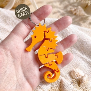 May include: Orange seahorse keychain with a silver keyring. The seahorse is segmented and has a detailed design. The keychain is held in a hand. The text "QUICK & EASY" is in a circle in the upper left corner.