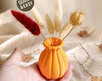 May include: A small orange pumpkin-shaped vase with a fluted rim containing dried botanical stems, including one deep red plume, three cream-colored fluffy grass stalks, and one round tan seed pod. A circular badge reads QUICK & EASY.