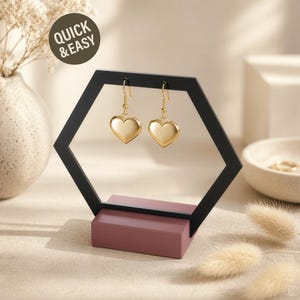 Puede incluir: Un expositor de pendientes hexagonal negro con una base malva, que sostiene dos pendientes dorados en forma de corazón. Los pendientes cuelgan dentro del marco geométrico. El texto "QUICK & EASY" está en un círculo en el fondo.