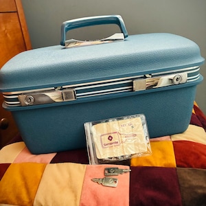 May include: A blue hardshell train case with a textured finish, a top handle, and chrome-coloured latches featuring keyholes. Included are two metal keys and a clear plastic pouch containing a paper insert with the text Samsonite and KEY 124.