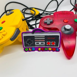 May include: A purple and black sticker with a retro video game controller design. The sticker says "Old School" and has the words "Select" and "Start" on the controller. The sticker is next to a red and a yellow Nintendo 64 controller.