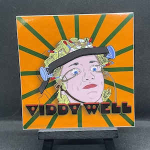 May include: A sticker featuring a cartoon illustration of a person with wires attached to their head. The background is orange with green stripes. The text "VIDDY WELL" is printed in black with red outlines.