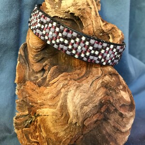 May include: A beaded bracelet with a black leather backing. The beads are in shades of purple, white, and silver.