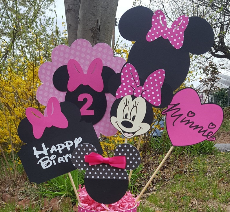 Puede incluir: Un conjunto de seis decoraciones de fiesta de Minnie Mouse en rosa y negro. Las decoraciones incluyen un letrero en forma de coraz&oacute;n que dice "Minnie", un letrero que dice "Feliz cumplea&ntilde;os", un letrero con el n&uacute;mero "2" y tres recortes de cabeza de Minnie Mouse.