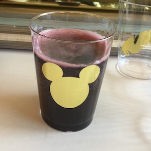 Puede incluir: Un vaso de pl&aacute;stico transparente con una bebida de color morado oscuro y una pegatina de Mickey Mouse dorada en la parte delantera.