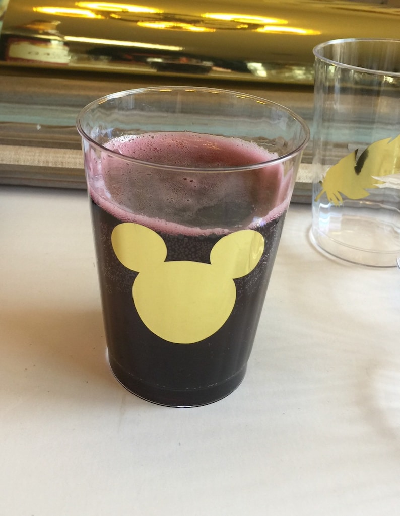 Puede incluir: Un vaso de pl&aacute;stico transparente con una bebida de color morado oscuro y una pegatina de Mickey Mouse dorada en la parte delantera.