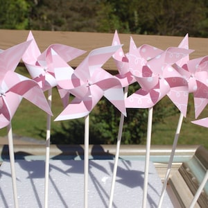 May include: A group of pink and white paper pinwheels with different patterns, including polka dots, stripes, and swirls. The pinwheels are attached to white sticks and are arranged in a row.