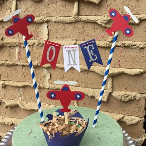 Puede incluir: Un adorno para pastel de avi&oacute;n rojo, blanco y azul con una pancarta que dice "ONE" en letras plateadas con purpurina. El adorno para pastel es perfecto para una fiesta de primer cumplea&ntilde;os.