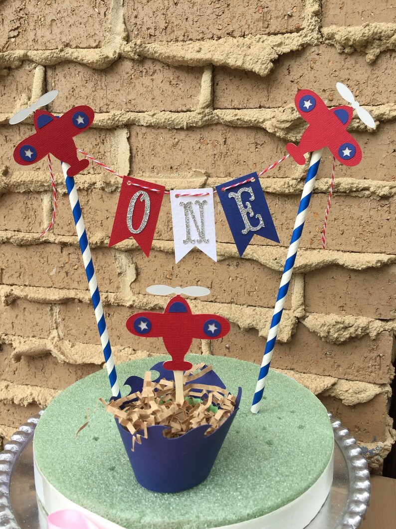 Puede incluir: Un adorno para pastel de avi&oacute;n rojo, blanco y azul con una pancarta que dice "ONE" en letras plateadas con purpurina. El adorno para pastel es perfecto para una fiesta de primer cumplea&ntilde;os.