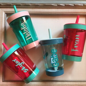 May include: Four personalized, colorful, plastic cups with straws and lids. The cups are teal, red, green, and blue. The names "Skylar", "Fran", "Gabe", and "Natalia" are written on the cups.