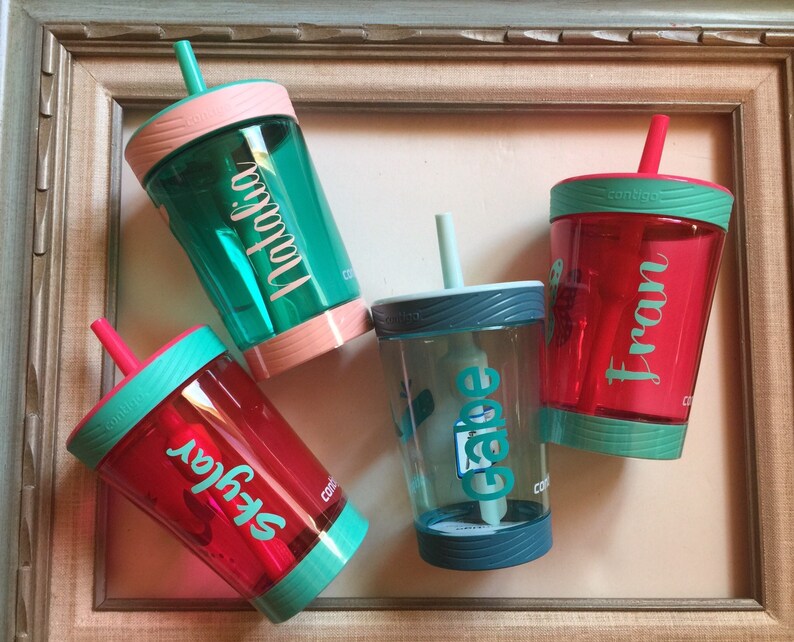 May include: Four personalized, colorful, plastic cups with straws and lids. The cups are teal, red, green, and blue. The names "Skylar", "Fran", "Gabe", and "Natalia" are written on the cups.