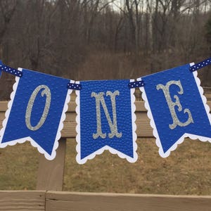 Puede incluir: Bandera azul y blanca con la palabra "ONE" en letras plateadas con purpurina. La bandera est&aacute; decorada con dos aviones azules y rojos.