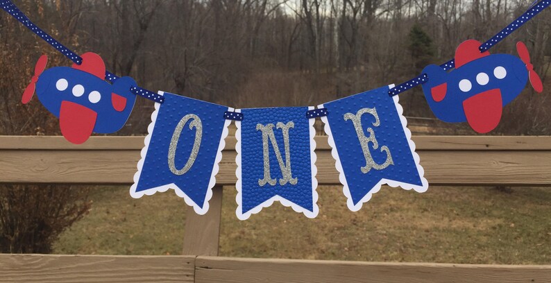 Puede incluir: Bandera azul y blanca con la palabra "ONE" en letras plateadas con purpurina. La bandera est&aacute; decorada con dos aviones azules y rojos.