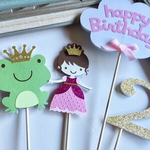 May include: A set of three cake toppers featuring a green frog, a princess in a pink dress, and a gold glitter number two. The princess has a crown and brown hair. The frog has a gold crown. The cloud topper says "happy birthday" in pink letters.