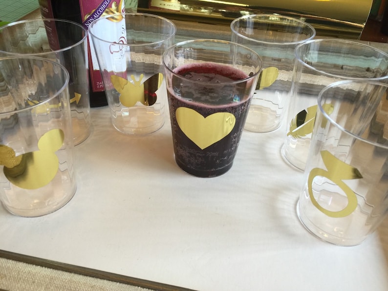 Puede incluir: Seis vasos de pl&aacute;stico transparentes con dise&ntilde;os de l&aacute;mina dorada. Los dise&ntilde;os incluyen un coraz&oacute;n, un p&aacute;jaro, una pluma, un anillo y una abeja. Un vaso contiene un l&iacute;quido rojo oscuro.