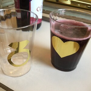 Puede incluir: Dos vasos de pl&aacute;stico transparentes con detalles en l&aacute;mina dorada. Un vaso tiene el n&uacute;mero "3" en dorado. El otro vaso tiene un coraz&oacute;n dorado y est&aacute; lleno de un l&iacute;quido rojo oscuro.