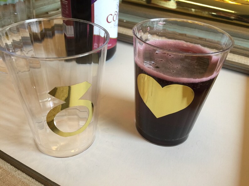 Puede incluir: Dos vasos de pl&aacute;stico transparentes con detalles en l&aacute;mina dorada. Un vaso tiene el n&uacute;mero "3" en dorado. El otro vaso tiene un coraz&oacute;n dorado y est&aacute; lleno de un l&iacute;quido rojo oscuro.