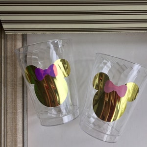 Puede incluir: Dos vasos de pl&aacute;stico transparentes con orejas de Mickey Mouse doradas y un lazo. Un lazo es morado y el otro es rosa.