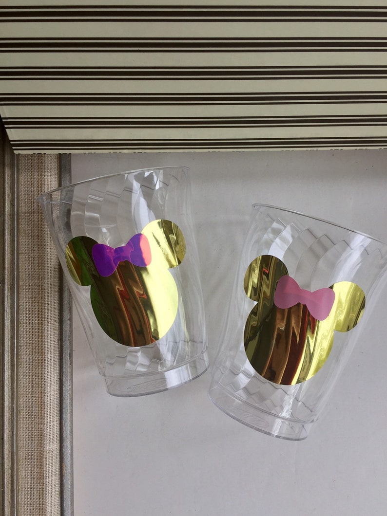 Puede incluir: Dos vasos de pl&aacute;stico transparentes con orejas de Mickey Mouse doradas y un lazo. Un lazo es morado y el otro es rosa.