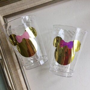 Puede incluir: Dos vasos de pl&aacute;stico transparentes con pegatinas doradas de la cabeza de Mickey Mouse. Un vaso tiene un lazo rosa y el otro un lazo morado.