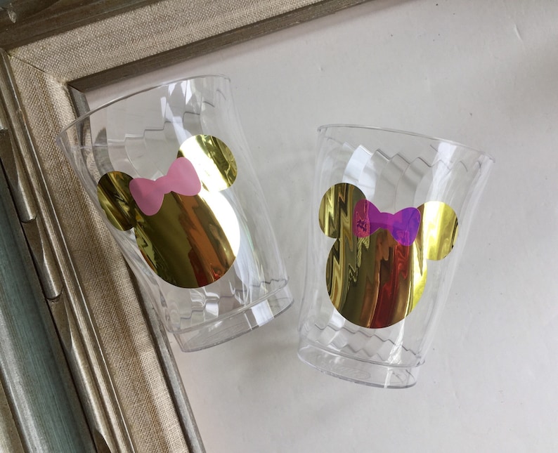 Puede incluir: Dos vasos de pl&aacute;stico transparentes con pegatinas doradas de la cabeza de Mickey Mouse. Un vaso tiene un lazo rosa y el otro un lazo morado.