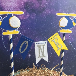 Puede incluir: Toppers de pastel de helic&oacute;ptero azul y amarillo con una pancarta blanca que dice "ONE" en letras plateadas brillantes.