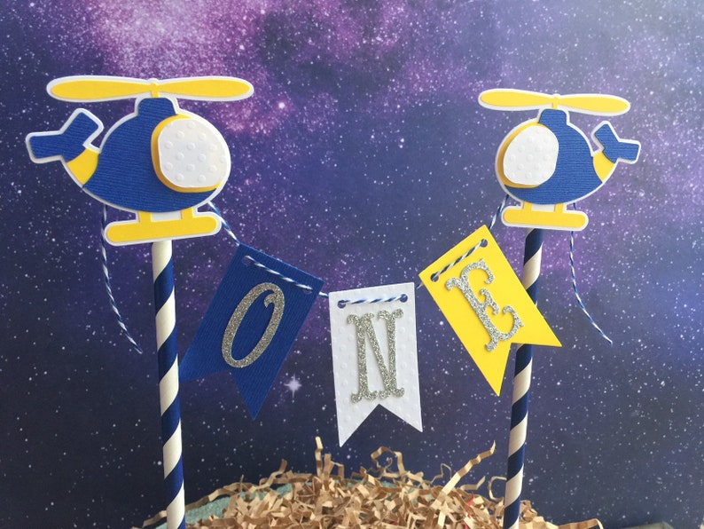 Puede incluir: Toppers de pastel de helic&oacute;ptero azul y amarillo con una pancarta blanca que dice "ONE" en letras plateadas brillantes.