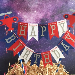 Puede incluir: Topper de pastel rojo, blanco y azul con una pancarta que dice "Feliz cumplea&ntilde;os" y dos aviones a cada lado.