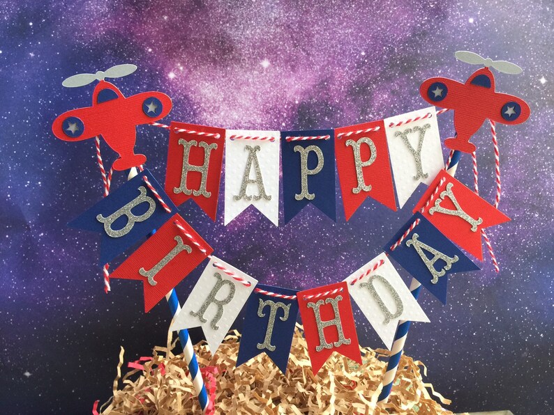 Puede incluir: Topper de pastel rojo, blanco y azul con una pancarta que dice "Feliz cumplea&ntilde;os" y dos aviones a cada lado.