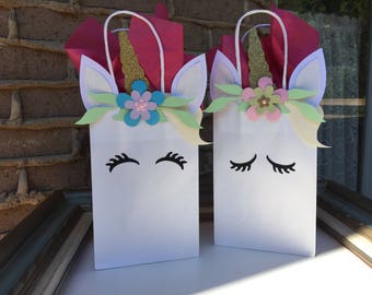 May include: Two white unicorn-themed gift bags with gold glitter horns, pink, blue, and green flowers, and black eyelashes. The bags have white handles.
